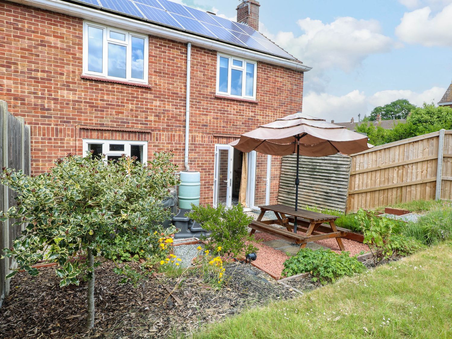 A backyard garden with a wooden picnic table and umbrella beside a brick house with solar panels at 3 Birkbeck Way in Norwich