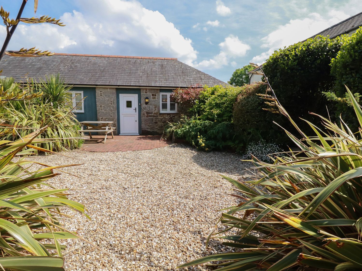A gravel courtyard with plants and shrubs surrounding a stone building with a white door and window at The Copper House in Bodrean near Truro