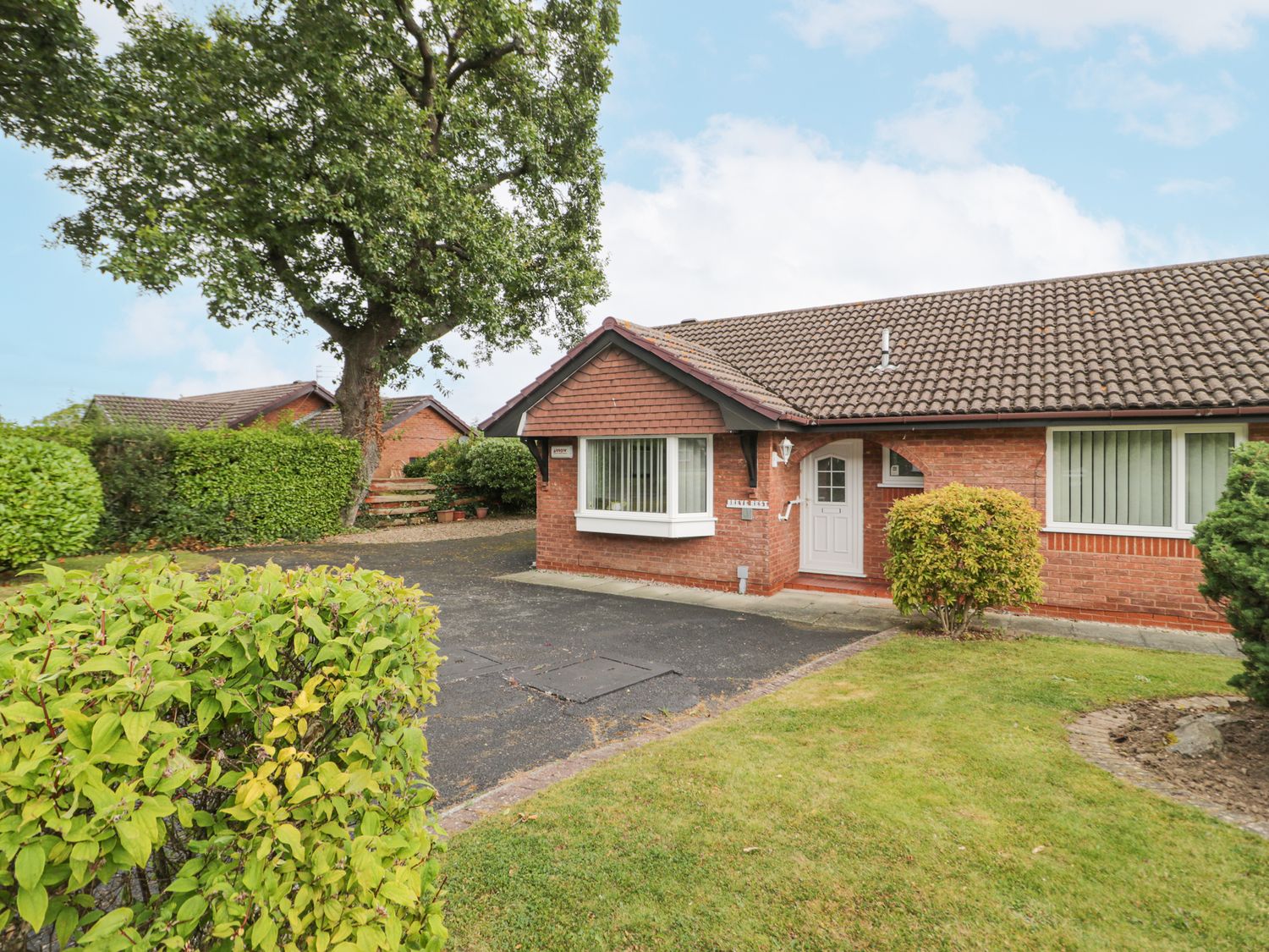 A single-story brick house with a tiled roof a white front door a driveway and green bushes at Breve Rest in Abergele