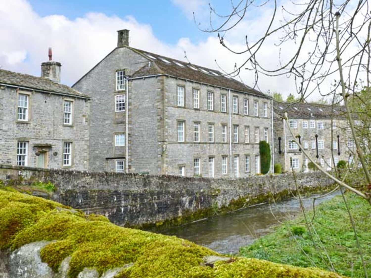 A row of stone buildings beside a narrow river with moss on the stone wall and trees at Riverside Walk in Airton near Malham
