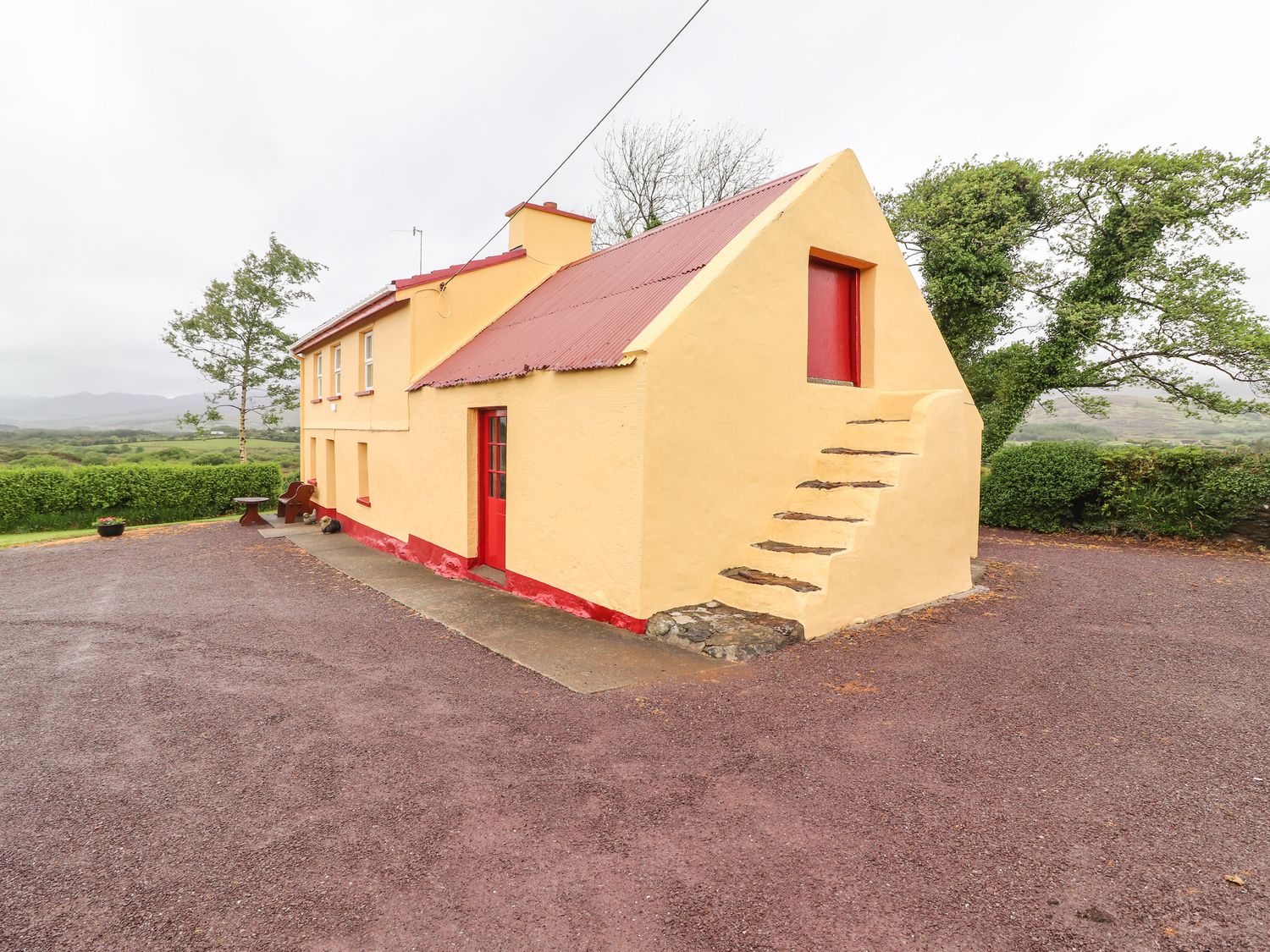 A yellow house with red doors and a red roof surrounded by trees and gravel at Ceol Na N'ean in Sneem County Kerry