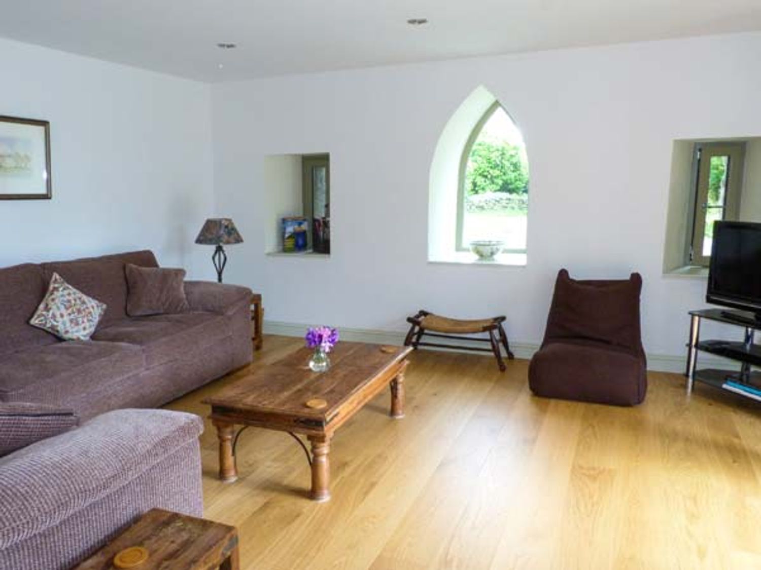 A living room with brown sofas a wooden coffee table a small stool a TV on a stand and arched windows at Muirmailing Cottage in Plean near Stirling