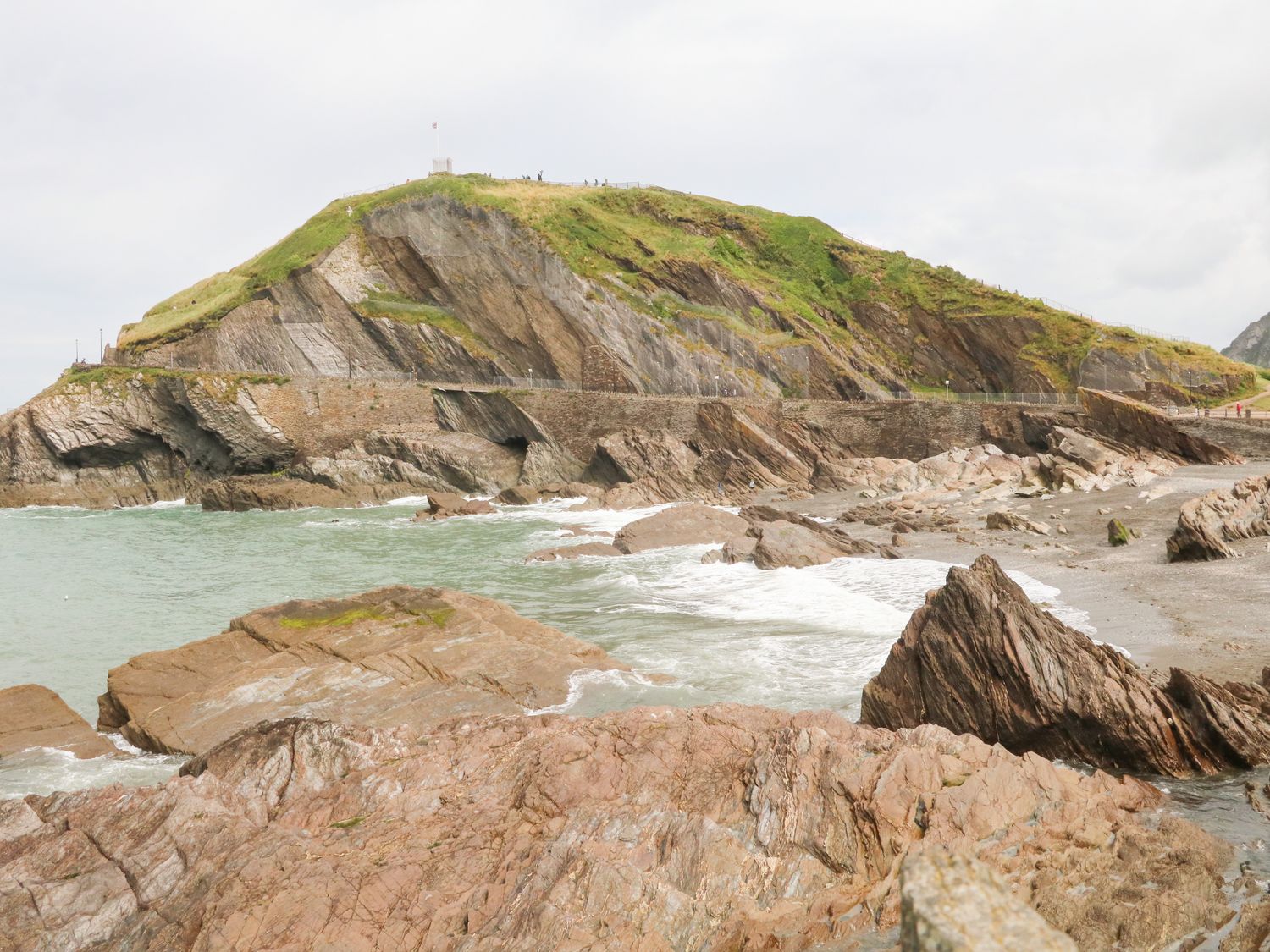 Rocky shoreline with a hill covered in grass and a stone wall near the water at Penryn in Ilfracombe