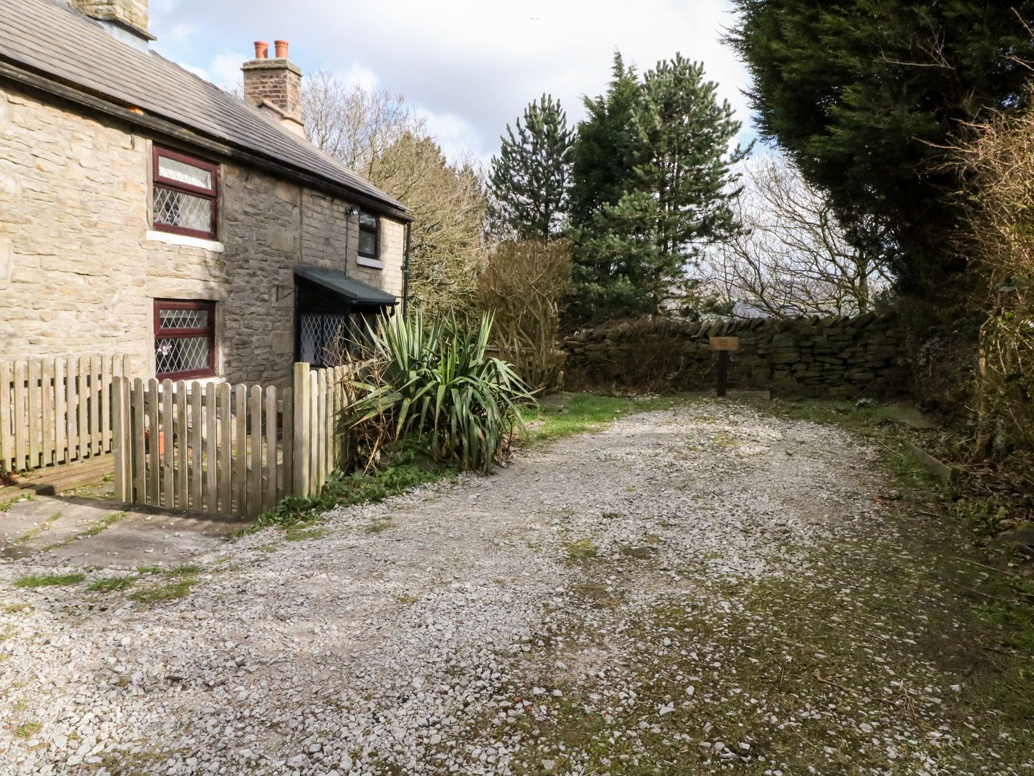 A stone house with a wooden gate and gravel driveway surrounded by trees and bushes at The Cottage Glossop in Charlesworth near Glossop