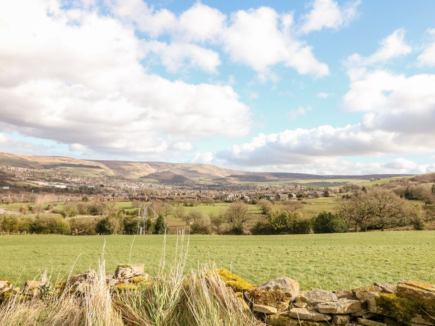 A rural landscape with green fields, a stone wall, trees, houses in the distance, and hills under a partly cloudy sky at The Cottage Glossop in Charlesworth near Glossop