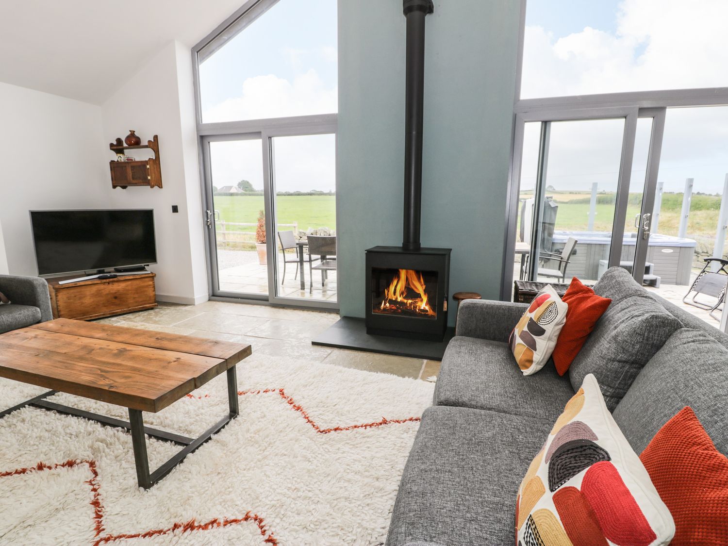 A living room with a gray sofa and colorful cushions a wooden coffee table a television and a fireplace with sliding glass doors showing a green field at Cae Sam in Pentraeth