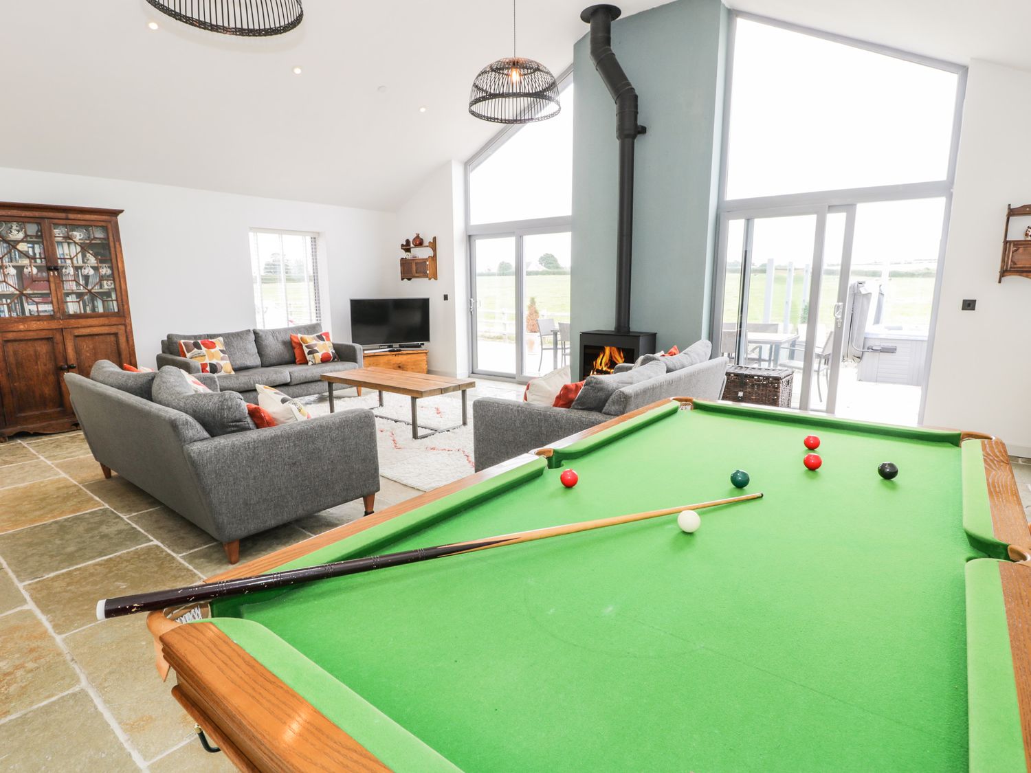 A living room with a pool table gray sofas a wood stove and large windows at Cae Sam in Pentraeth