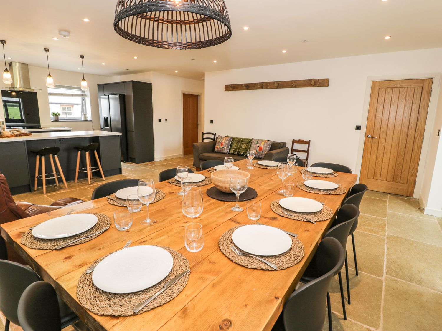 A dining room with wooden table set with plates glasses and cutlery and an open kitchen in the background at Cae Sam in Pentraeth