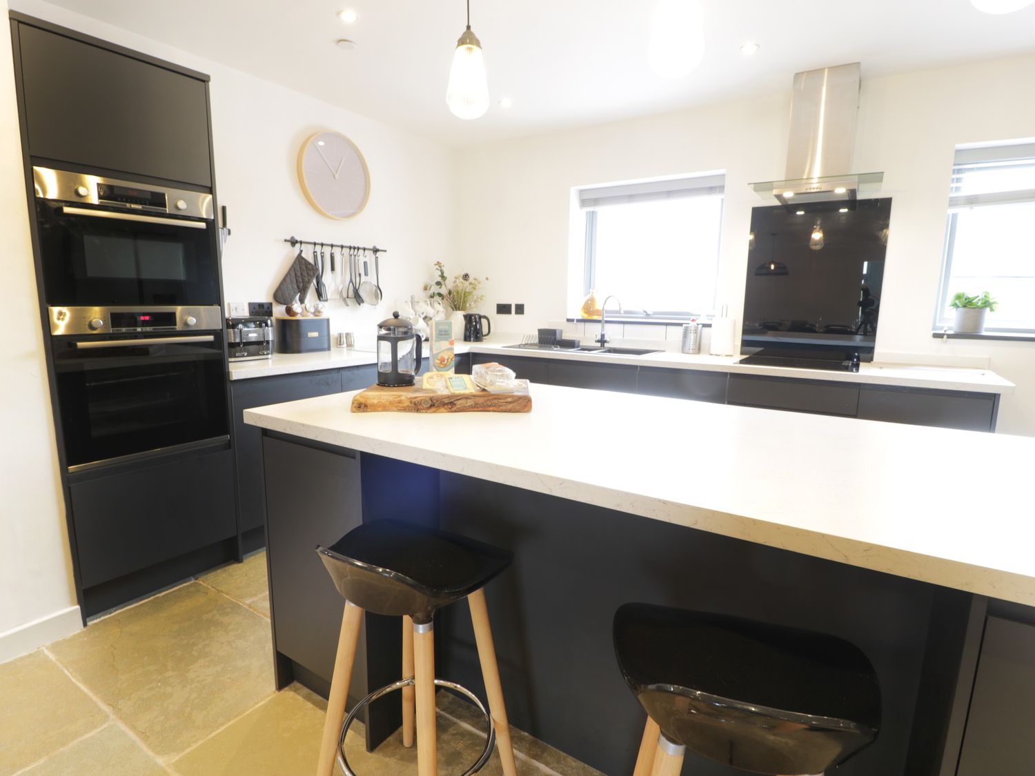A modern kitchen with black cabinets a white countertop two black stools wall clock and kitchen appliances at Cae Sam in Pentraeth