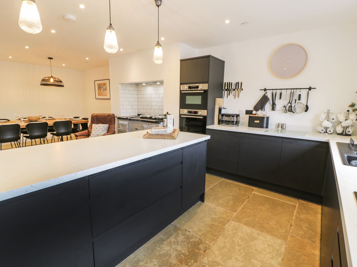 A kitchen with black cabinets white countertops a dining table with black chairs and a brown armchair at Cae Sam in Pentraeth
