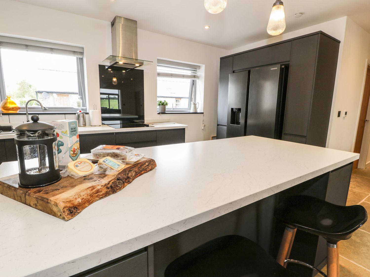 A modern kitchen with a white island countertop black stools black cabinets and a wooden cutting board with food items at Cae Sam in Pentraeth
