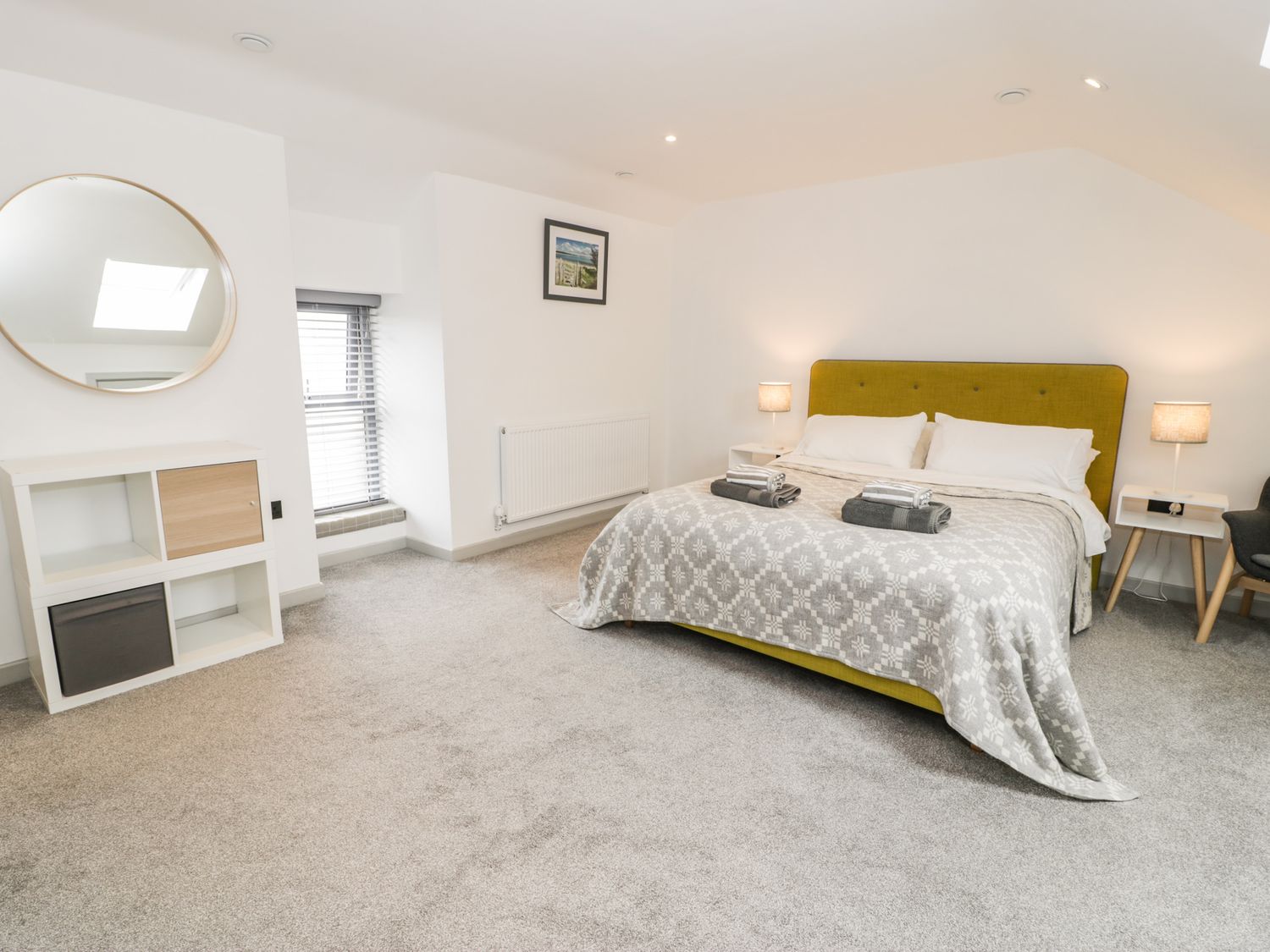 A bedroom with a double bed with a patterned blanket and two folded towel sets two bedside tables with lamps a round mirror on the wall a storage unit and a window with blinds at Cae Sam in Pentraeth