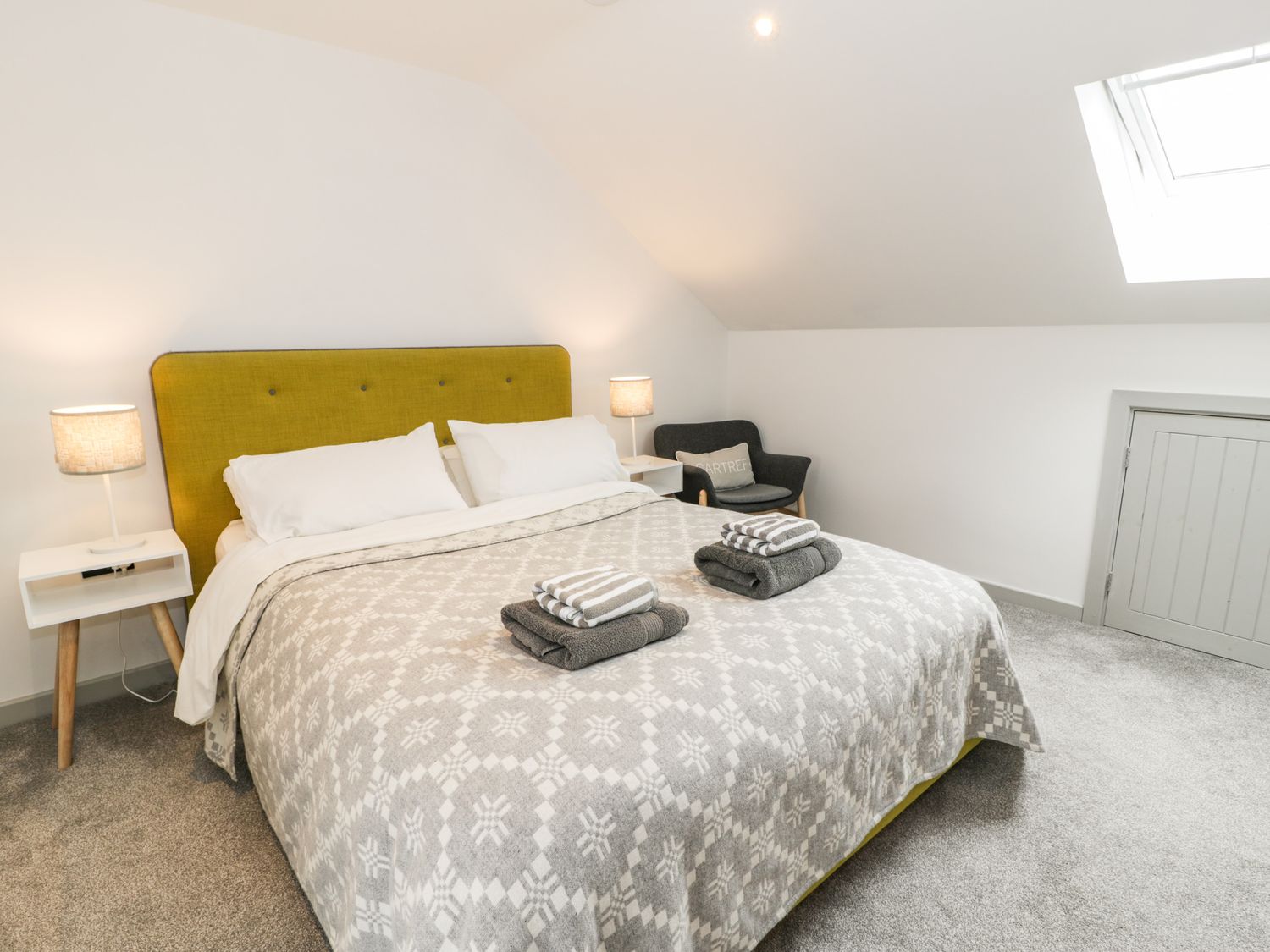 A bedroom with a bed with a yellow headboard two bedside tables with lamps a chair a skylight window and a small door at Cae Sam in Pentraeth