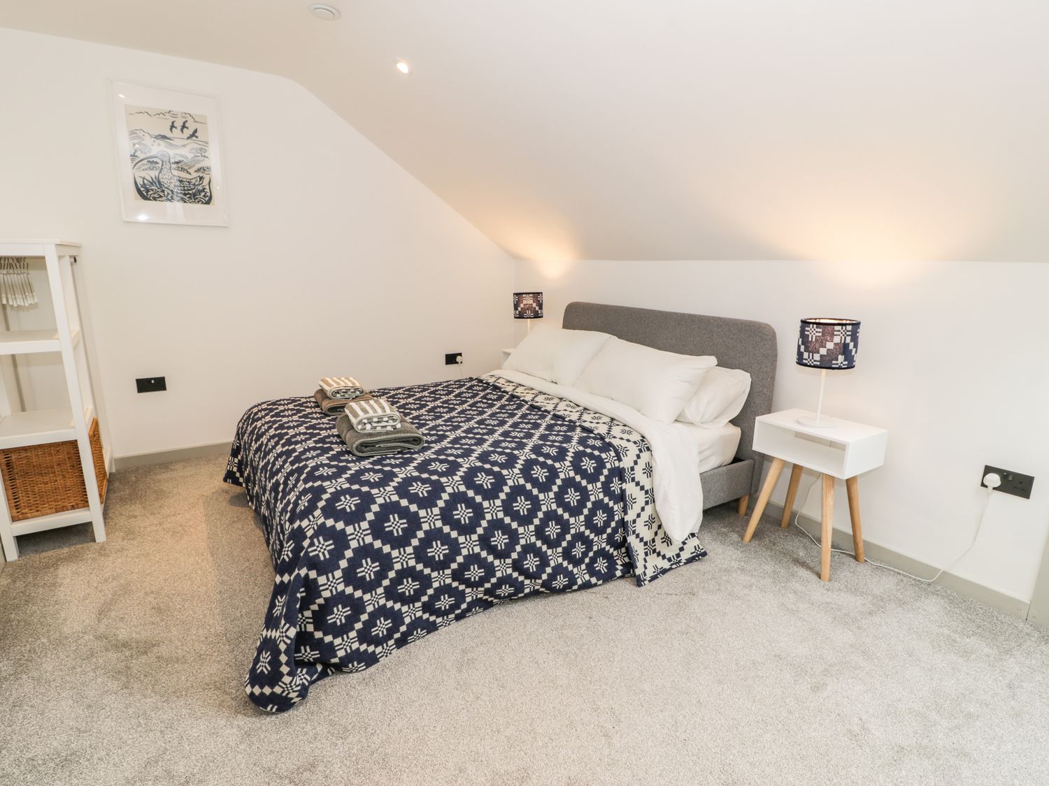 A bedroom with a bed with patterned blanket two bedside tables with lamps and a shelving unit with baskets at Cae Sam in Pentraeth
