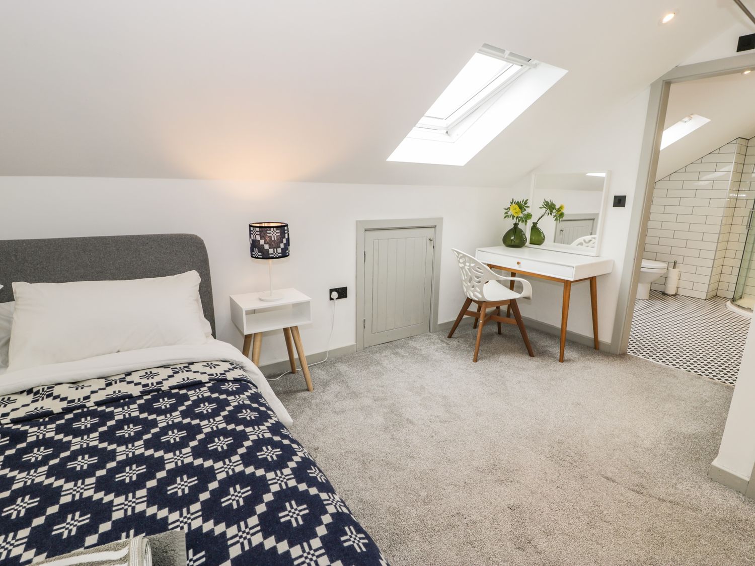 A bedroom with a bed a bedside table with a lamp a small door a desk with a chair and a skylight window in a room with carpet at Cae Sam in Pentraeth