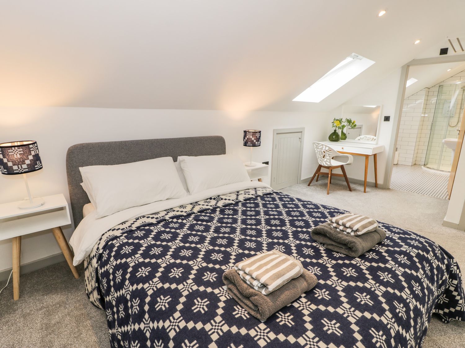 A bedroom with a bed covered with a patterned blue and white blanket and towels on top a desk with a chair and a mirror and a skylight at Cae Sam in Pentraeth