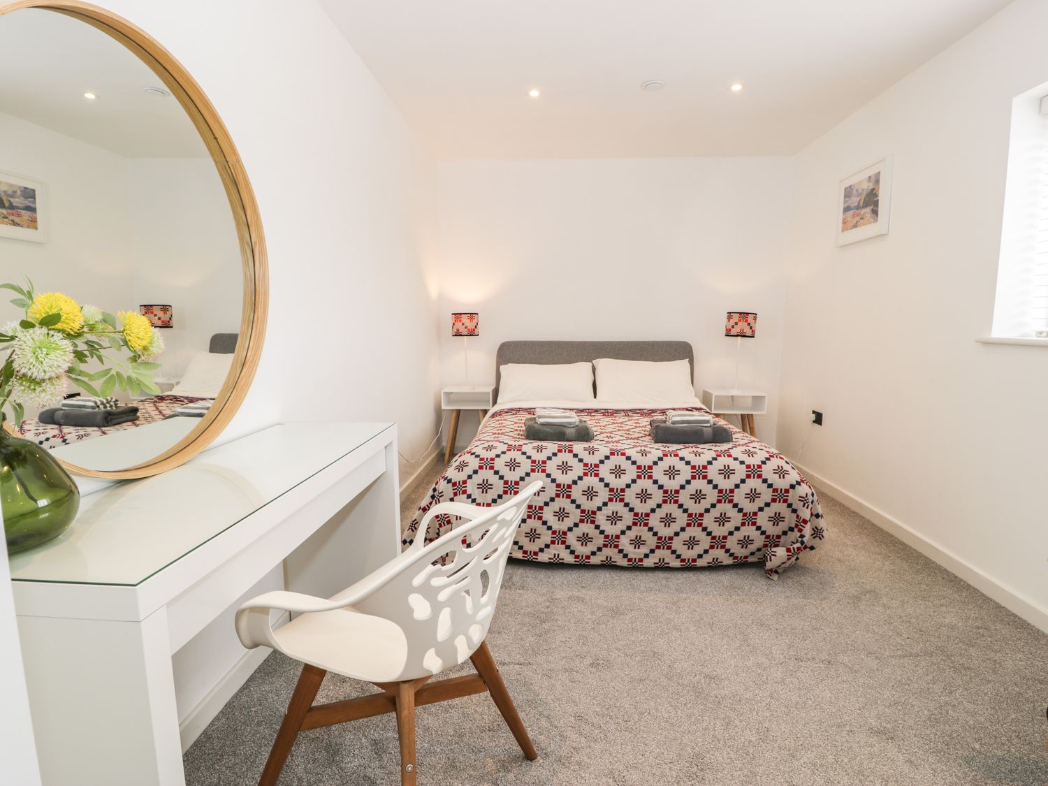 A bedroom with a double bed two bedside tables lamps a white desk with a round mirror a white chair and a window at Cae Sam in Pentraeth