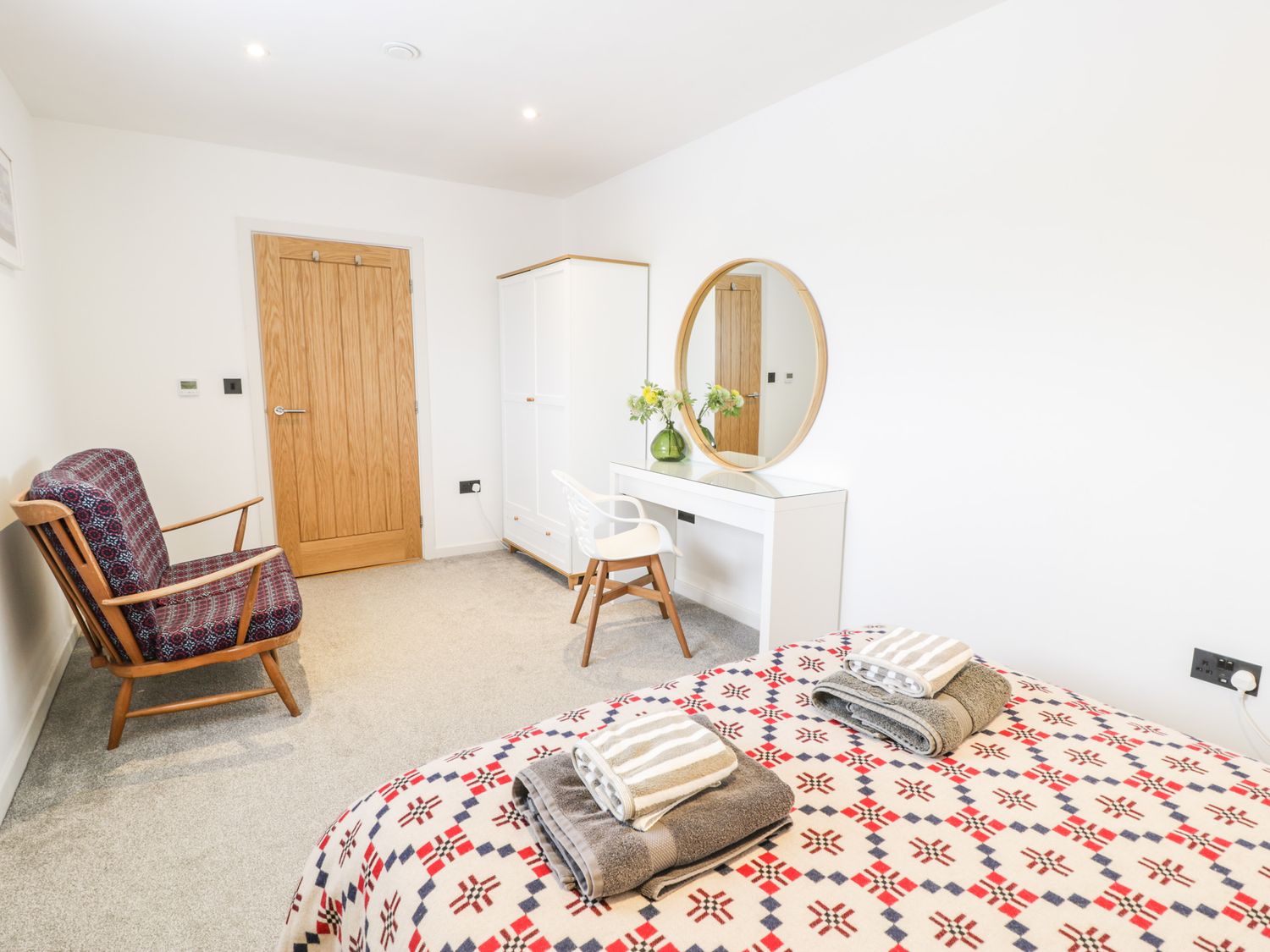 A bedroom with patterned bedspread folded towels on the bed wooden armchair white wardrobe desk with round mirror and vase of flowers at Cae Sam in Pentraeth