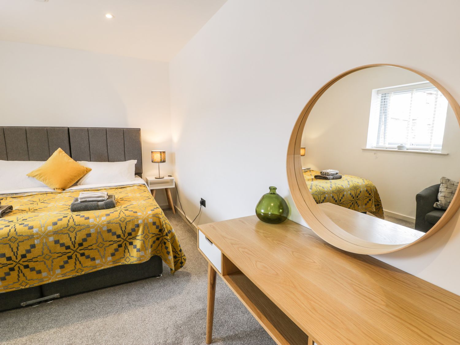 A bedroom with a bed with a yellow patterned blanket a bedside table with a lamp a wooden dresser with a round mirror and a green vase at Cae Sam in Pentraeth