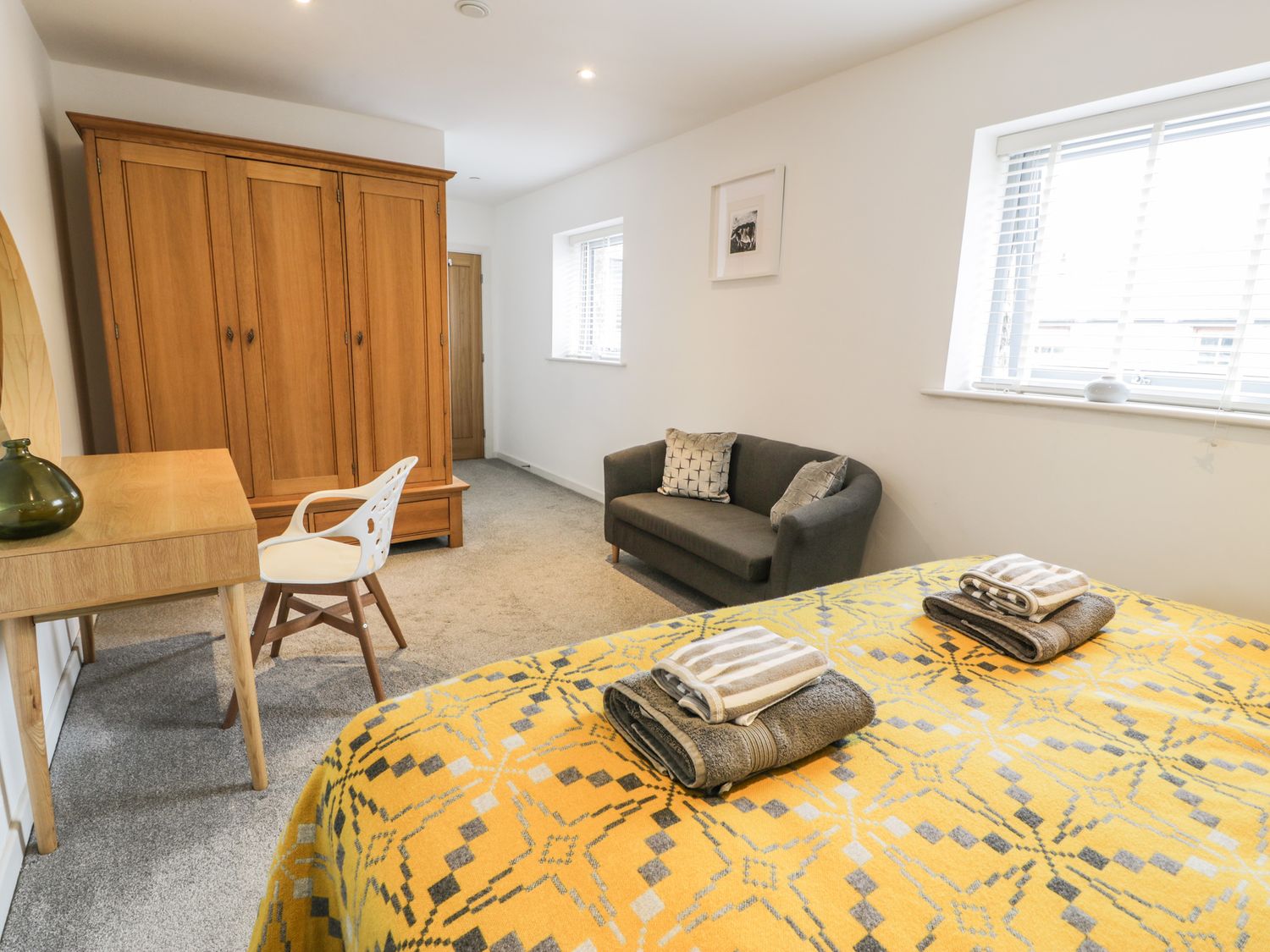 A bedroom with a yellow patterned bedspread, a wooden wardrobe, a small desk with a white chair, and a gray sofa at Cae Sam in Pentraeth