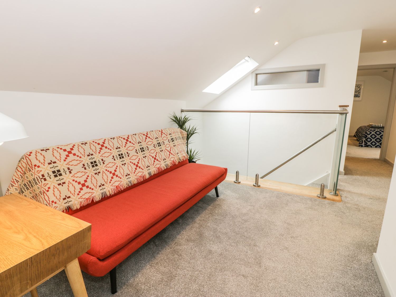 A sitting area with a red sofa covered with a patterned throw next to a wooden table near a glass railing at Cae Sam in Pentraeth