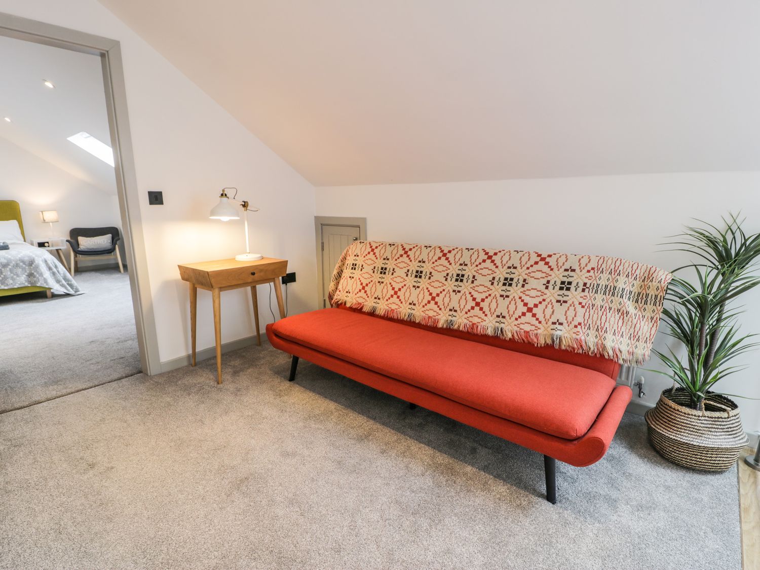 A room with an orange sofa covered with a patterned blanket next to a wooden side table with a lamp and a plant in a basket at Cae Sam in Pentraeth