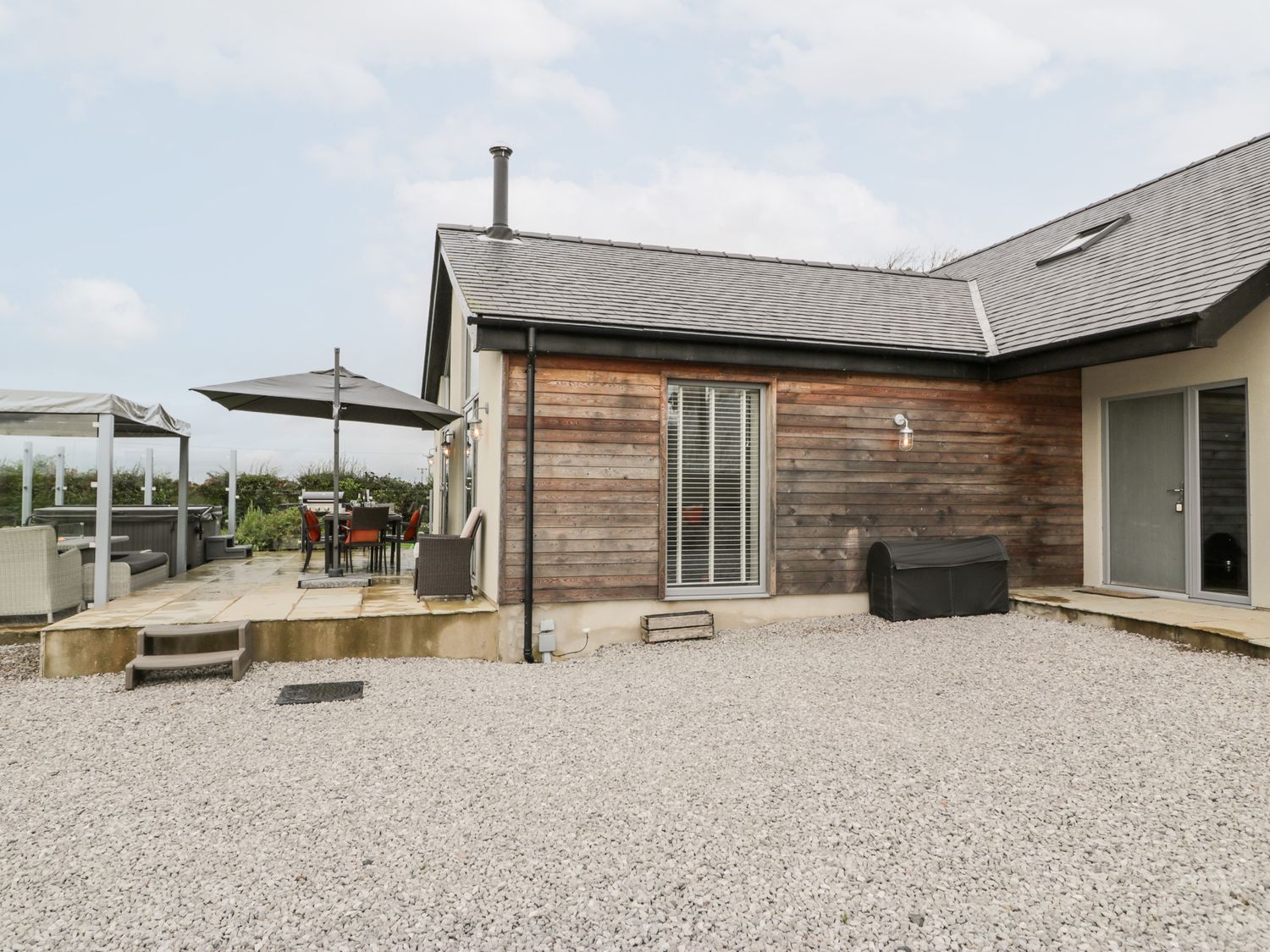 An outdoor patio area with gravel ground stones a wooden wall with a window and a door a covered grill a dining table with chairs under an umbrella and outdoor seating at Cae Sam in Pentraeth