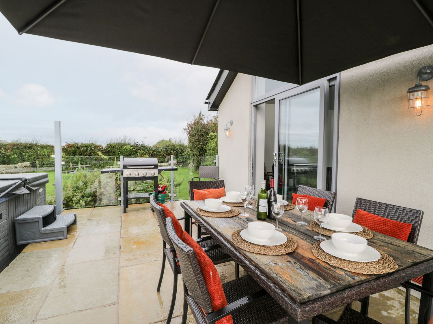 An outdoor patio with a dining table set with plates bowls wine glasses and bottles under an umbrella near a barbecue grill and hot tub at Cae Sam in Pentraeth