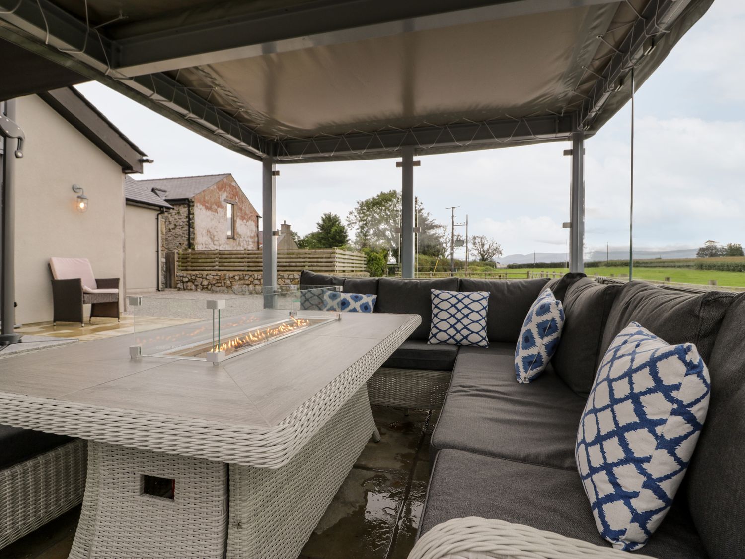 An outdoor seating area with a wicker sectional sofa and a rectangular fire table under a canopy at Cae Sam in Pentraeth