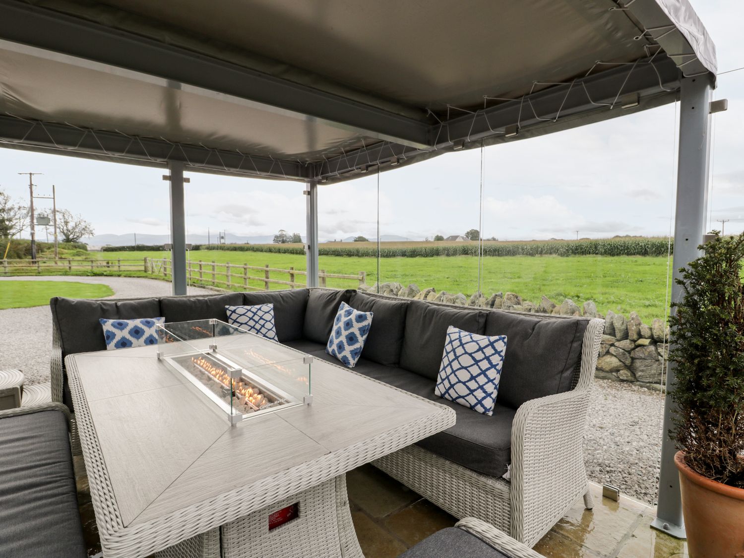 A covered outdoor seating area with a table featuring a built-in fire pit and a grey cushioned wicker sofa at Cae Sam in Pentraeth