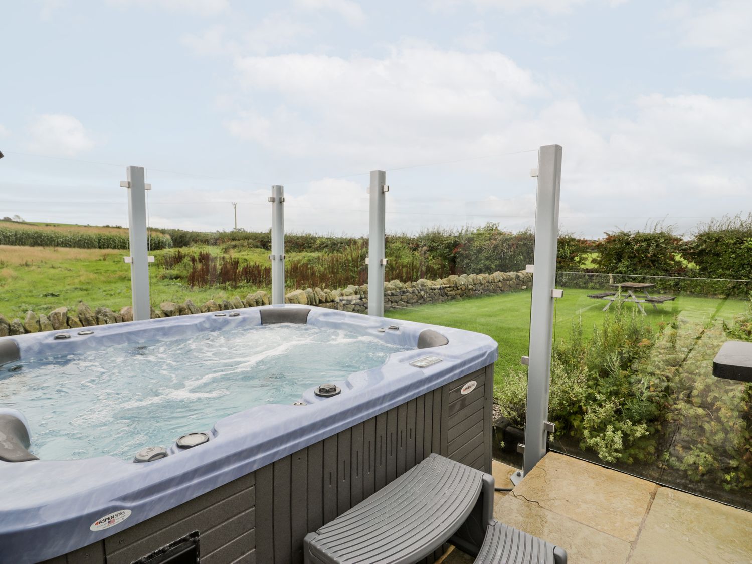 An outdoor hot tub with bubbling water on a patio with glass panel fencing and a grassy field with a picnic table beyond at Cae Sam in Pentraeth