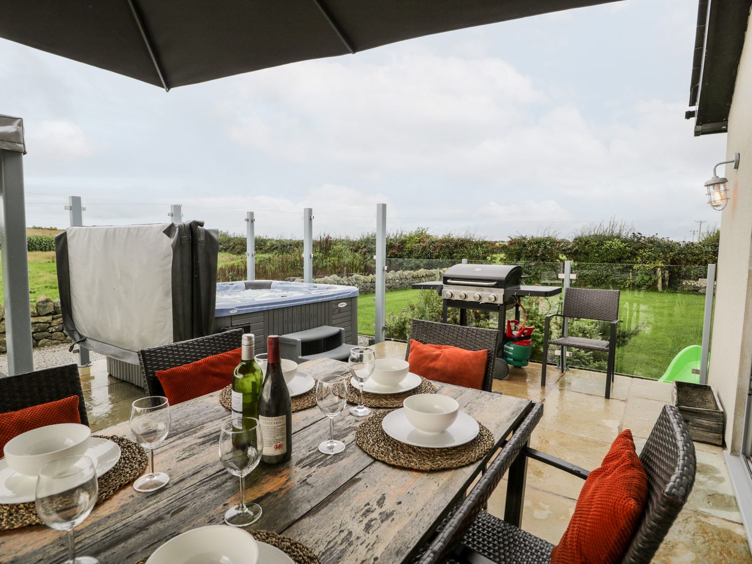 An outdoor patio with a dining table set with plates and wine glasses a barbecue a hot tub and chairs at Cae Sam in Pentraeth