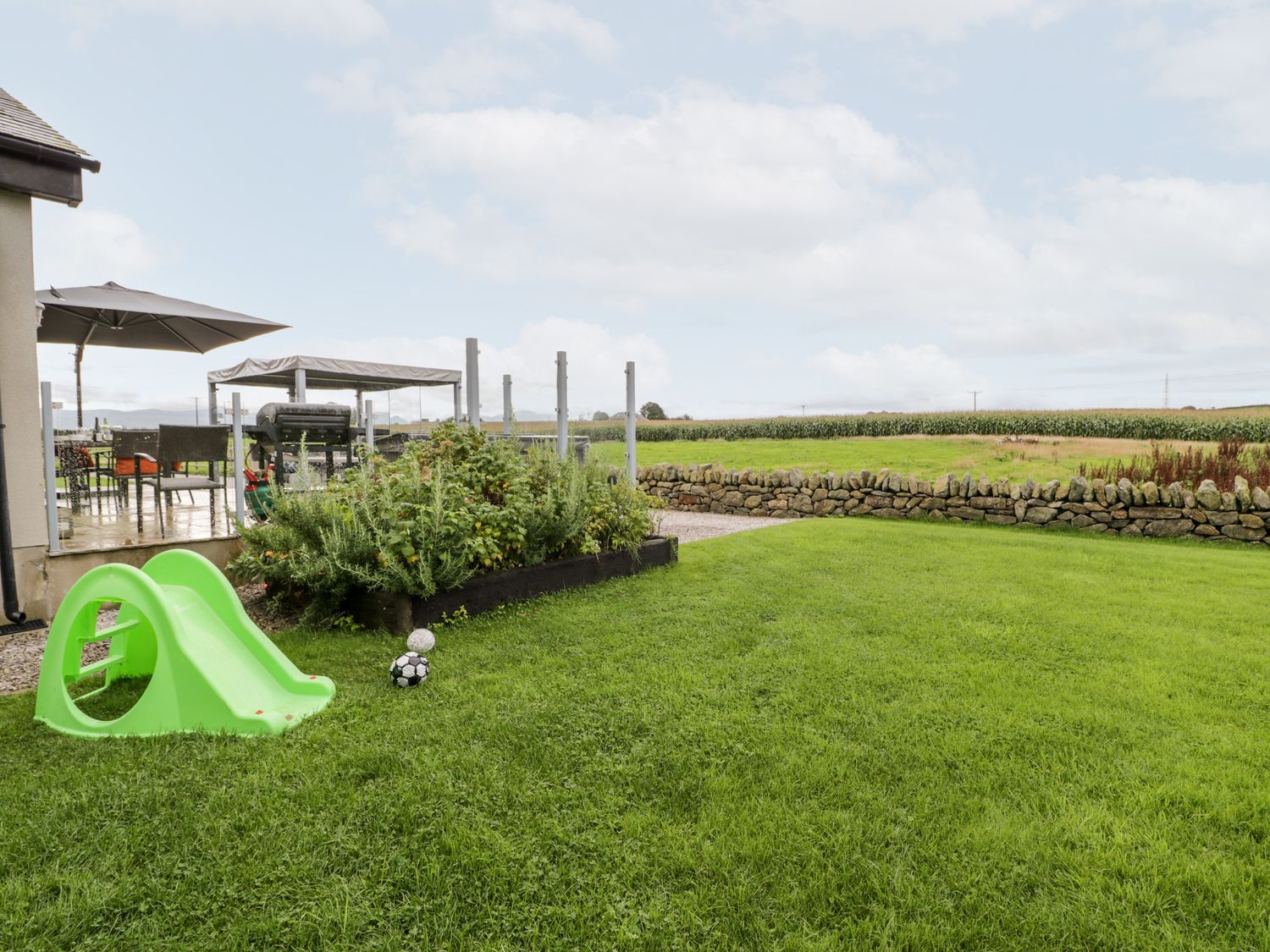 A backyard with green grass a small slide two balls a stone wall and a patio area at Cae Sam in Pentraeth