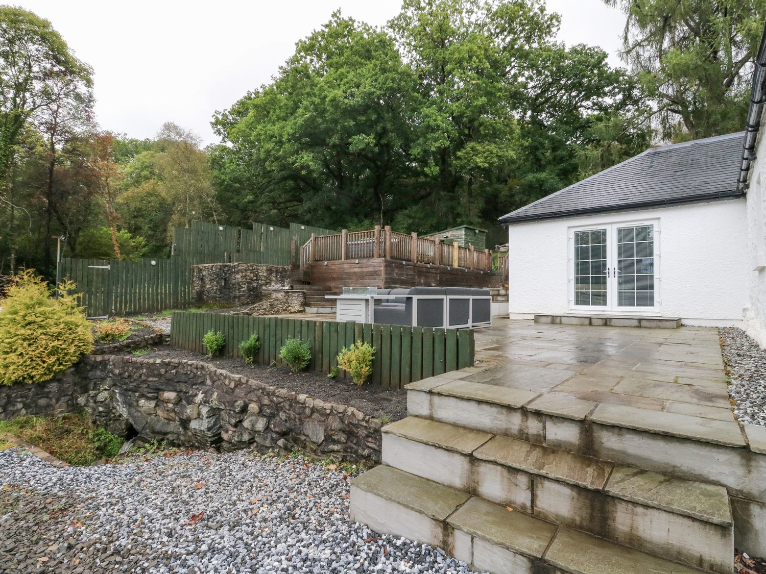 An outdoor patio area with stone steps stone retaining walls garden beds patio furniture and a white building with double doors at Heatherbank in Garelochhead