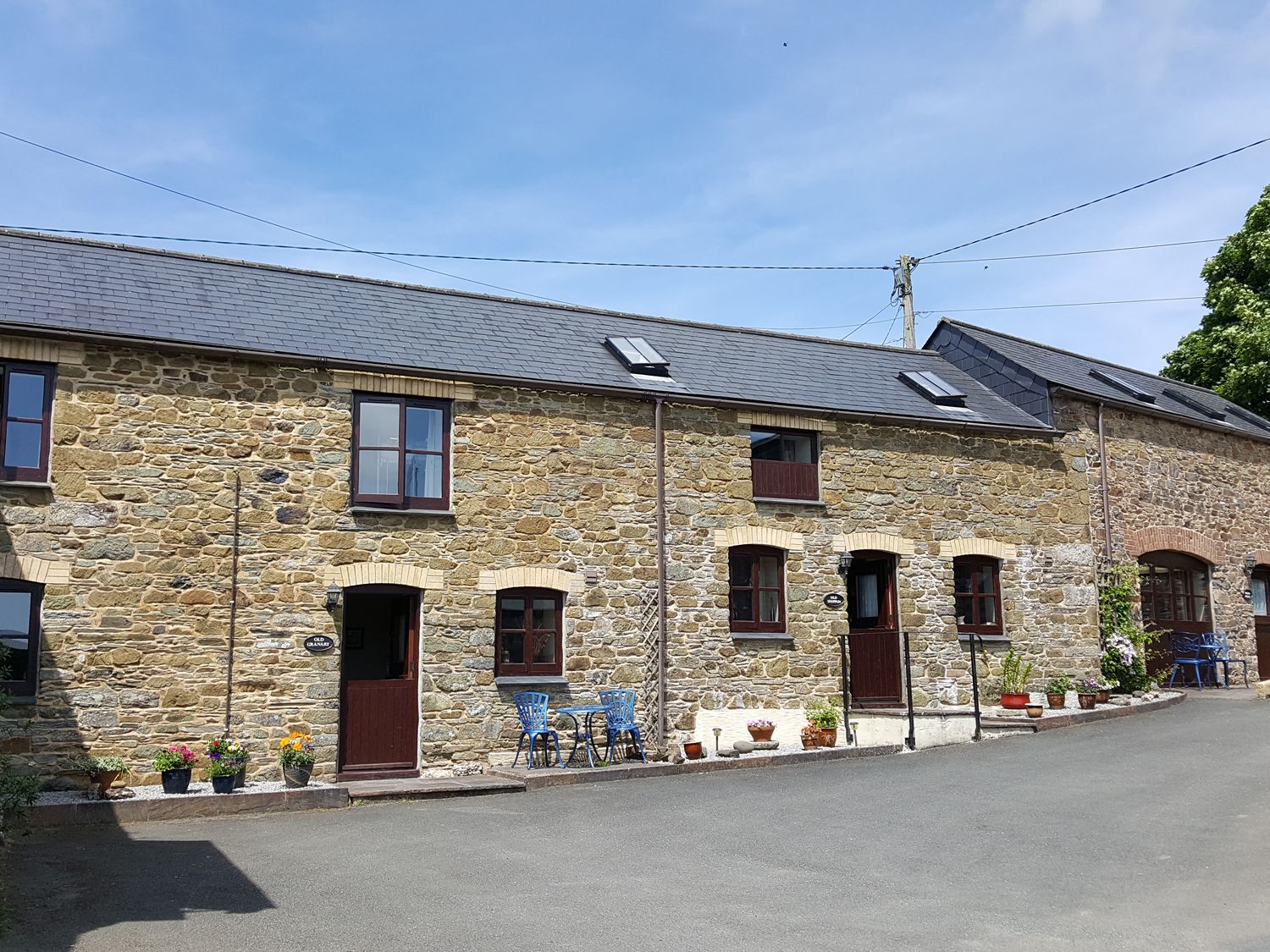 Stone cottages with wooden doors and windows with flower pots and blue chairs outside on a paved area at The Shippon Near Launceston