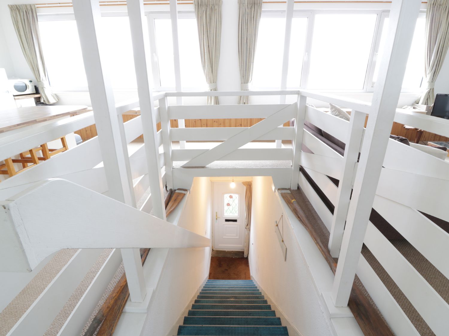 A staircase with blue carpet leading down to a white door in a room with white railings and windows at Top Flat in Tighnabruaich