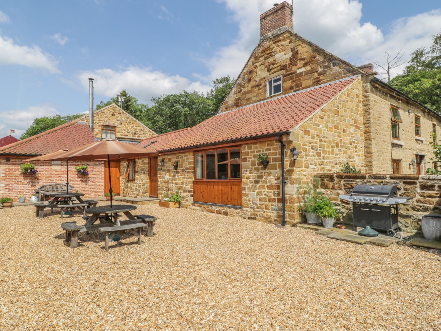 An outdoor area with stone buildings picnic tables umbrellas and a barbecue grill at Kitchen Cottage in Thirsk