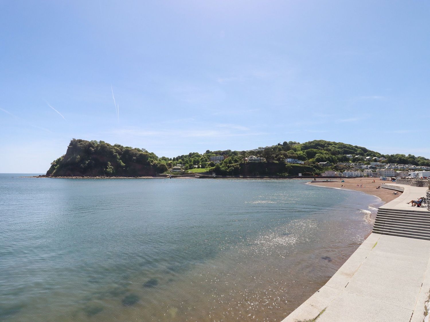 A curved beach with calm water a concrete promenade and green hills with houses in the background at Dunholme House in Teignmouth