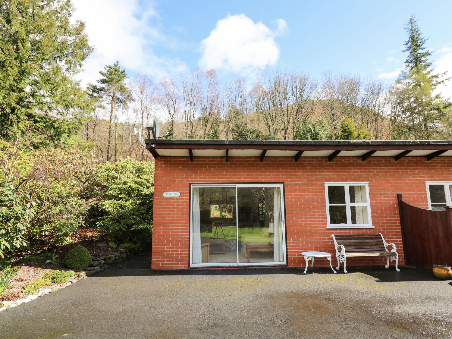 A brick building with sliding glass door bench and table in front surrounded by trees at Trout Cottage in Llanwrthwl near Rhayader