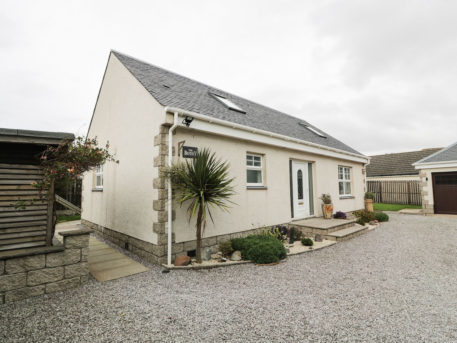 A single-story house with a white door and windows surrounded by gravel driveway and plants at The Bothy in Southerness