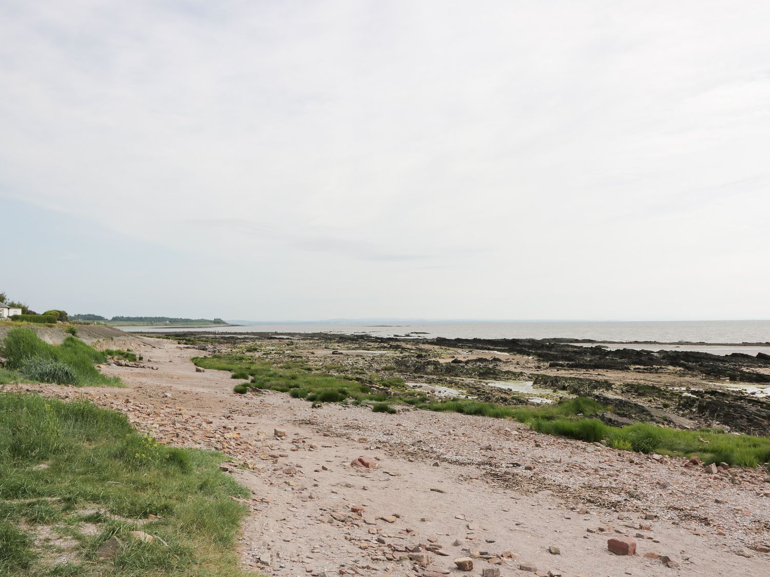 A rocky shoreline with patches of grass and a narrow dirt path by the sea at The Bothy in Southerness