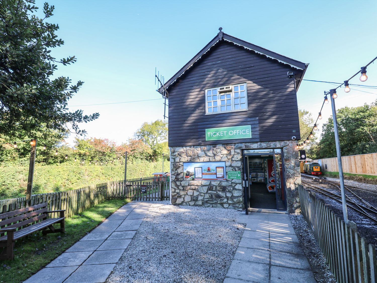 An outdoor ticket office building next to railway tracks with a bench and a fenced path at The Station Flat in St Newlyn East near Newquay