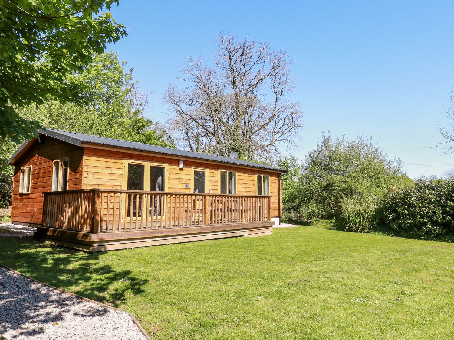 A wooden cabin with a deck on a grassy lawn surrounded by trees at Lodge One in Brighton near Summercourt