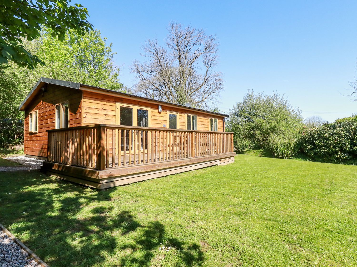 A wooden cabin with a deck and grass lawn surrounded by trees at Lodge One in Brighton near Summercourt