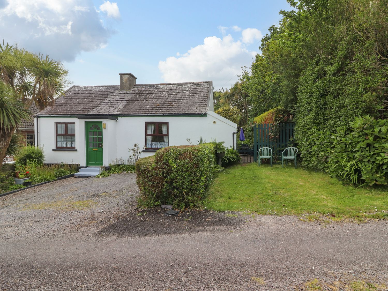 A white cottage with a green door and windows a gravel driveway and a garden with hedges and two plastic chairs at Brendans Cottage in Valentia Island County Kerry
