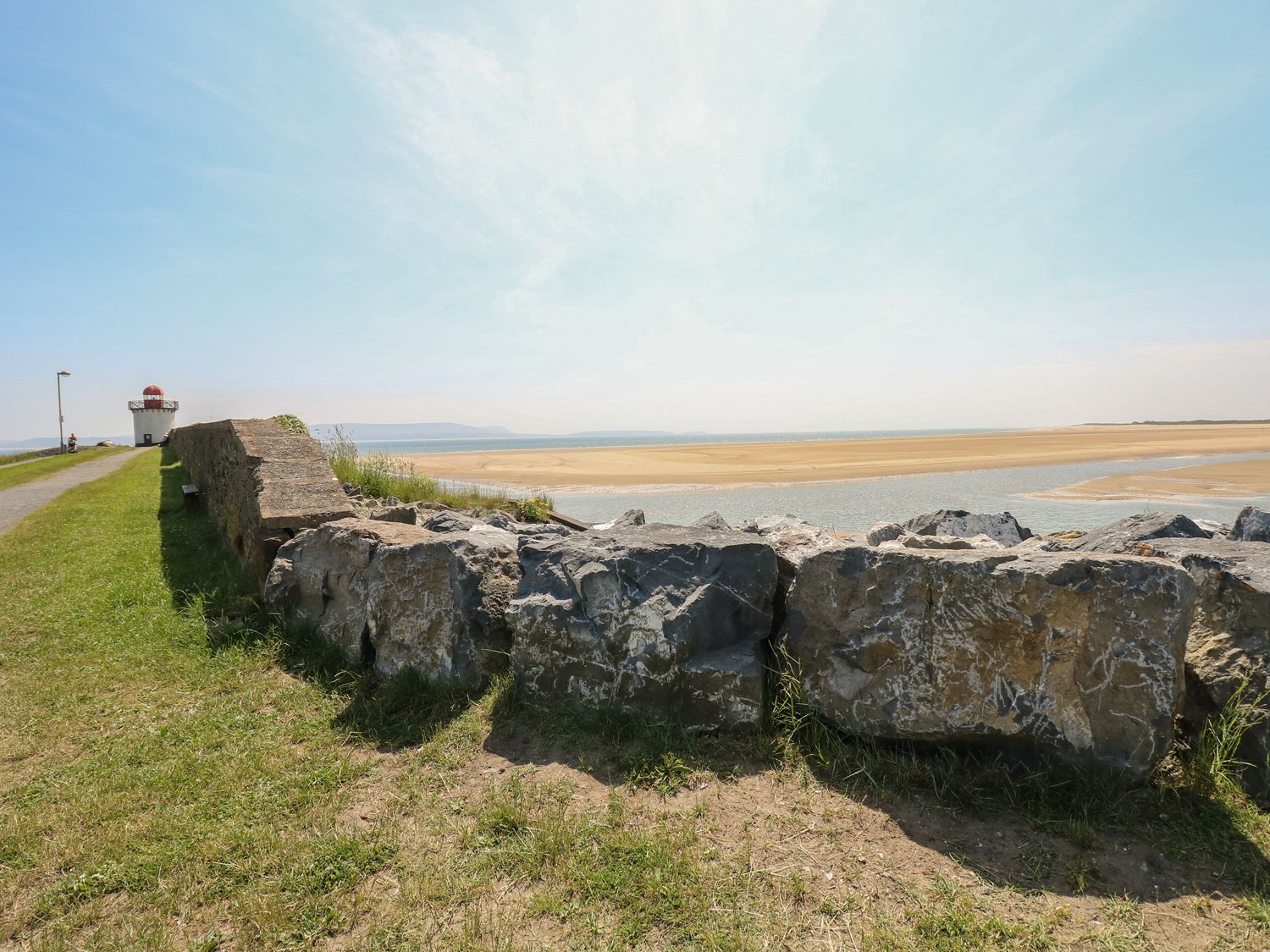 A grassy path with a stone wall leading to a small lighthouse near a sandy beach and water