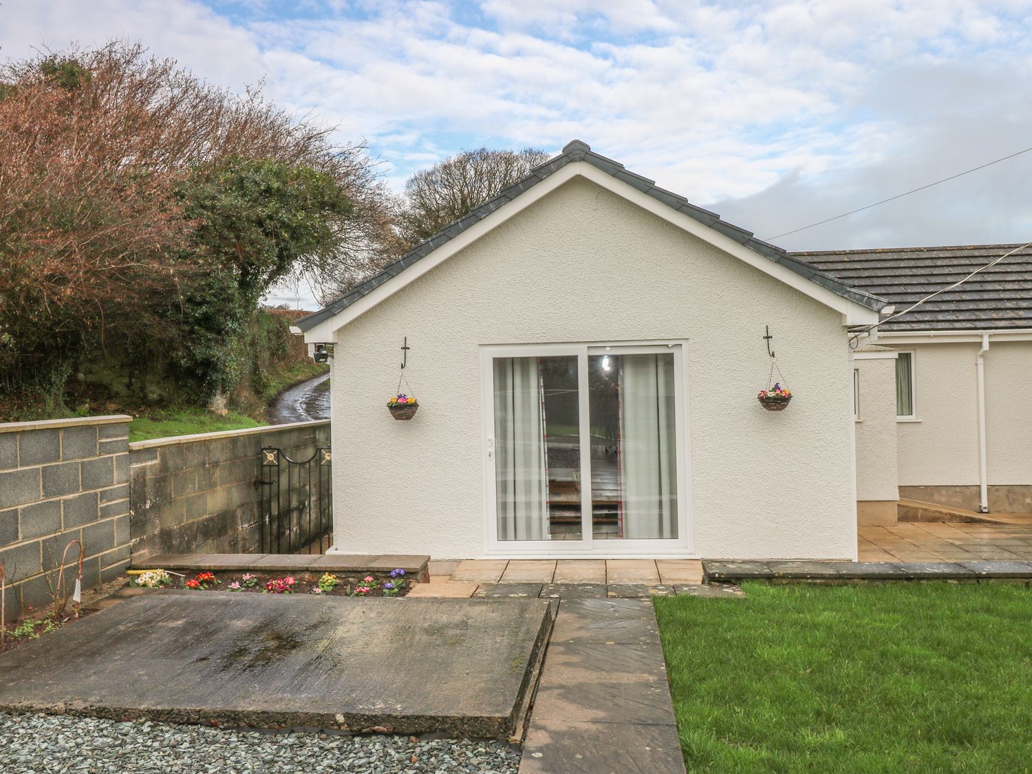 The back of a white bungalow with sliding glass doors two hanging flower baskets a concrete slab and a lawn at Broadford Farm Bungalow in Kidwelly