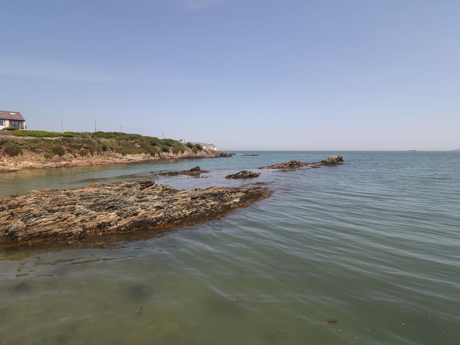 A rocky shoreline next to water with houses and vegetation in the background at Min Y Traeth in Bull Bay