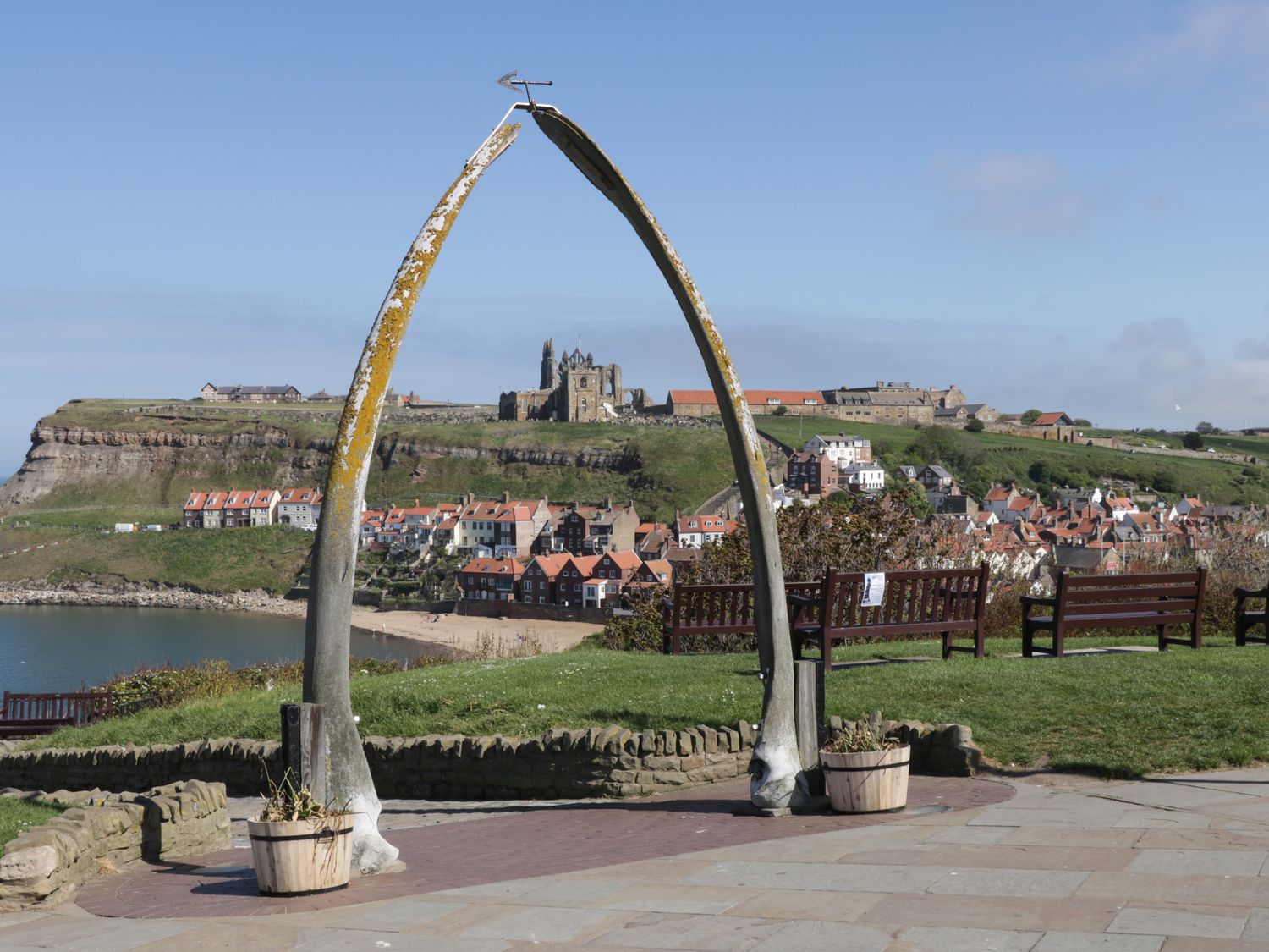 A coastal village with red roof houses and a historic abbey viewed through a large whale bone arch with benches on green grass near the shore at Barn Cottage in High Normanby near Robin Hood's Bay