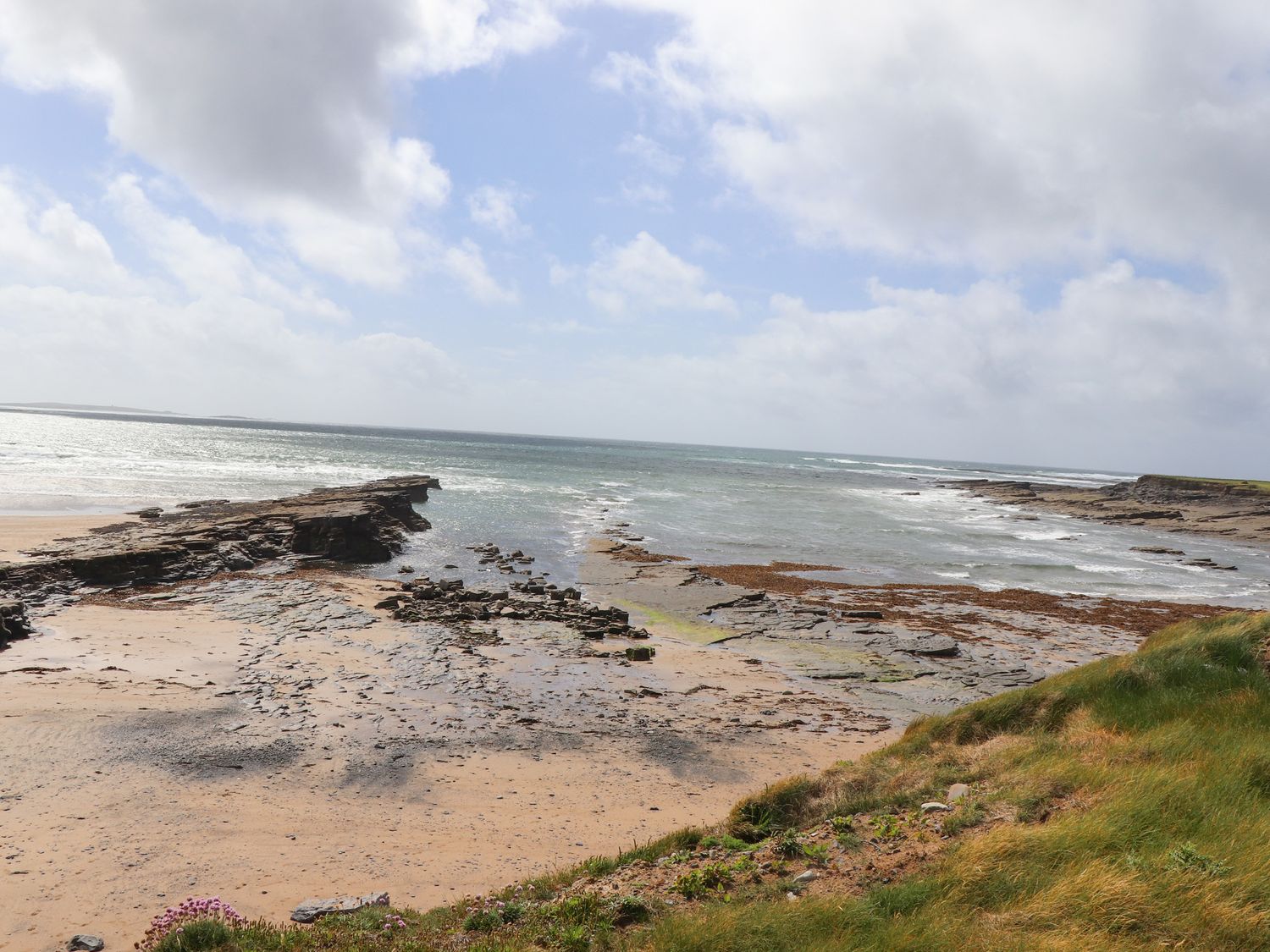 A rocky beach with sand and seaweed beside green grassy cliffs under a cloudy sky at Goodlands Cottage in Spanish Point County Clare